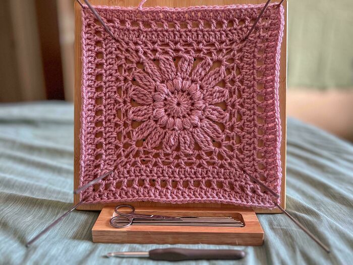 Pink crocheted square with floral pattern stretched on wooden frame, accompanied by crochet hooks and scissors nearby.
