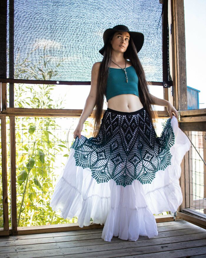 Woman wearing a black hat and a teal crop top showing a detailed crocheted black skirt overlay over a flowing white skirt.