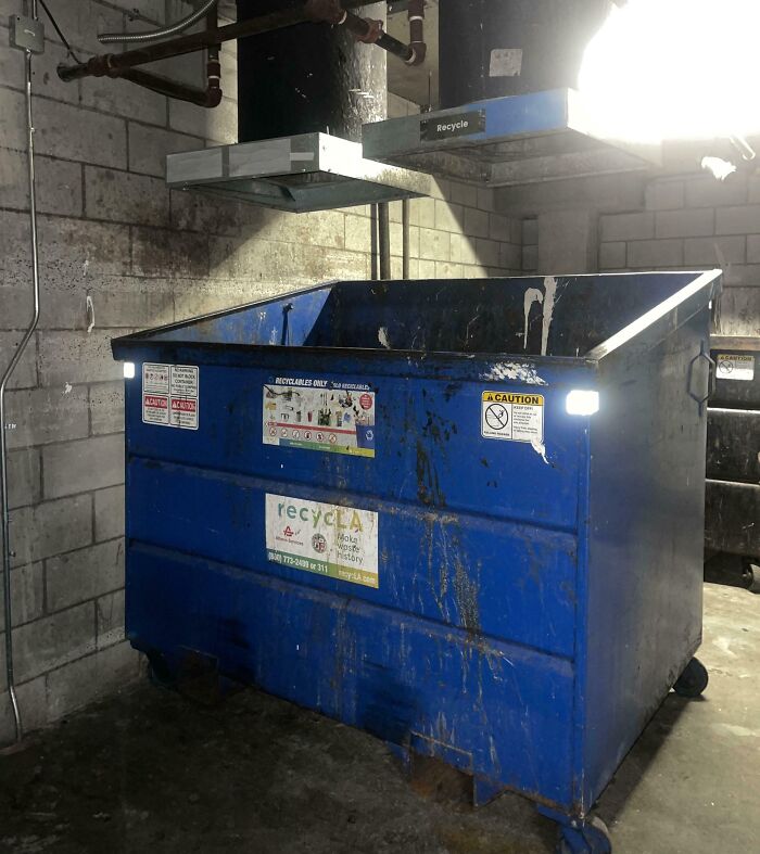 Blue industrial dumpster with recycling labels in a dimly lit, grimy concrete room, evoking dystopian US scene.
