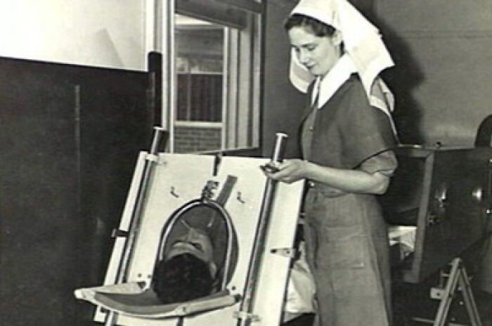 Vintage black and white medical pic showing a nurse operating an early treatment device in an old-school medical setting.
