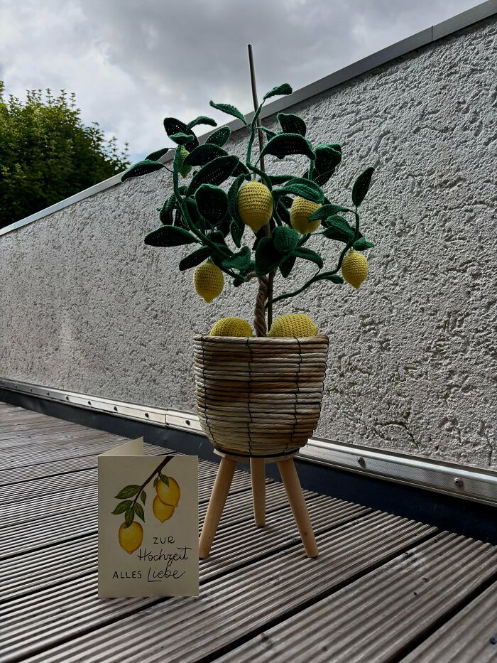 Crocheted lemon tree with green leaves in a woven pot on wooden stand displayed outdoors on decking.