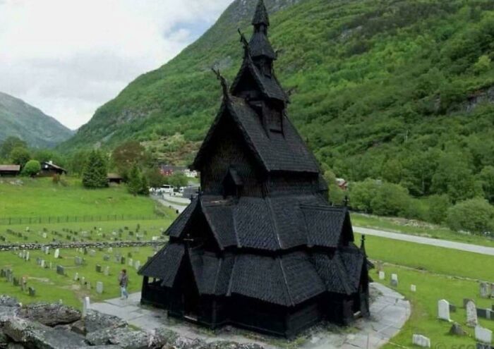 A creepy, dark wooden stave church with a towering spire, surrounded by a graveyard and lush green mountains.