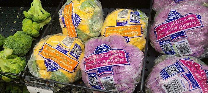 Packaged purple and orange cauliflower heads displayed among green broccoli, showcasing nonsensical real produce variations.