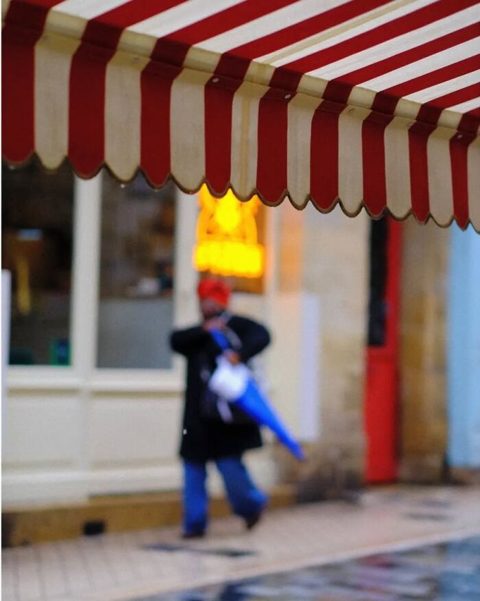 Blurred street scene with a person holding a blue umbrella under a red and white striped awning, street photos by Greek photographer.