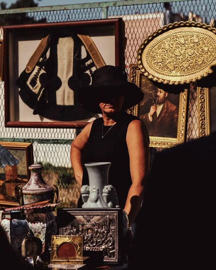 Street photo by Greek photographer Marina Nota showing a woman in a hat at an antique market stall.