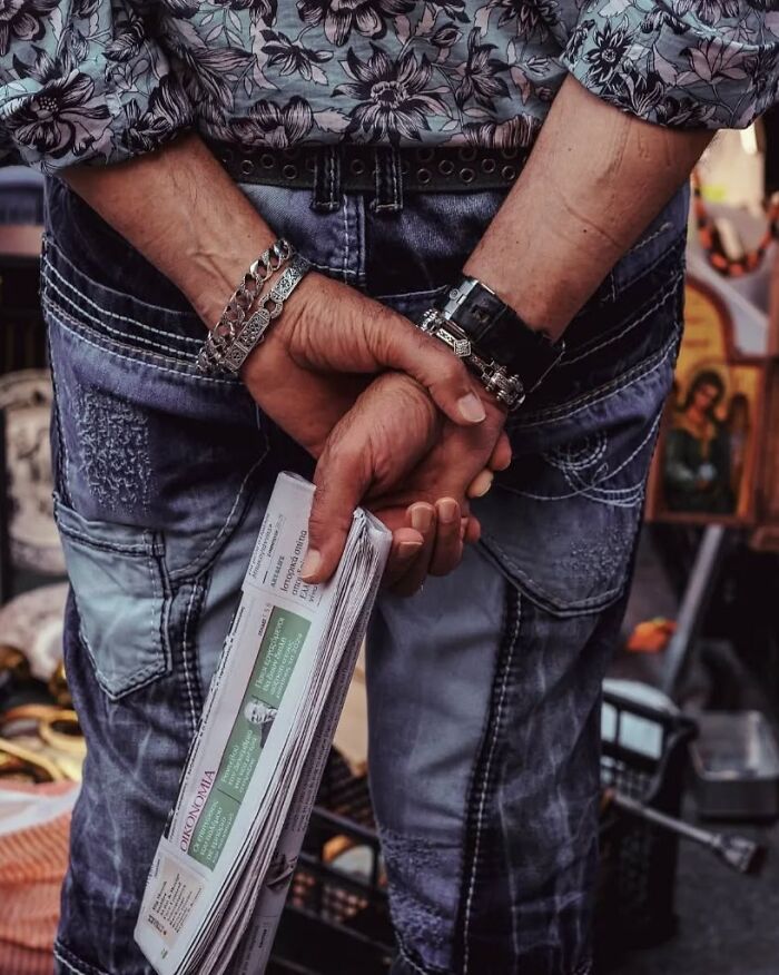 Close-up of a person holding a folded newspaper, wearing bracelets and patterned jeans in a street photo by Greek photographer.