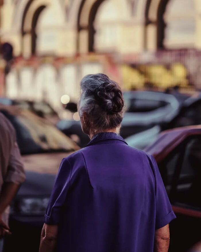 Elderly woman with gray hair in a bun wearing a purple coat in a busy urban street photo by Greek photographer.