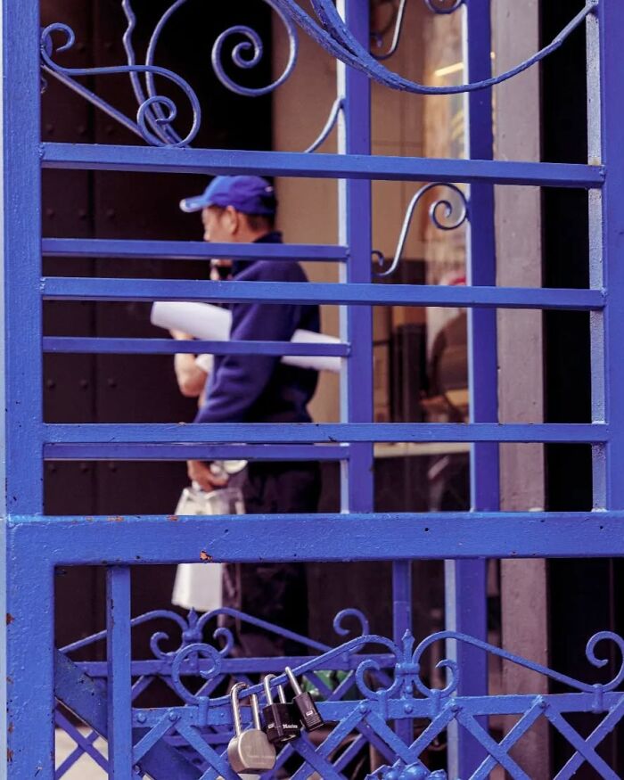 Blue metal gate with multiple locks in focus and a man holding rolled papers blurred in the background street photo.