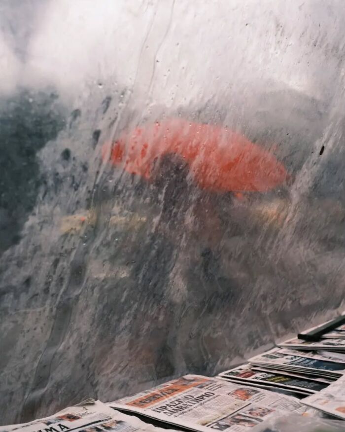 Blurred street scene with a person holding a red umbrella seen through a rain-covered window in street photos by Marina Nota.