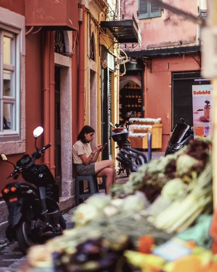 Young woman sitting on a stool in a narrow street with scooters and market produce in a Greek street photo by Marina Nota.