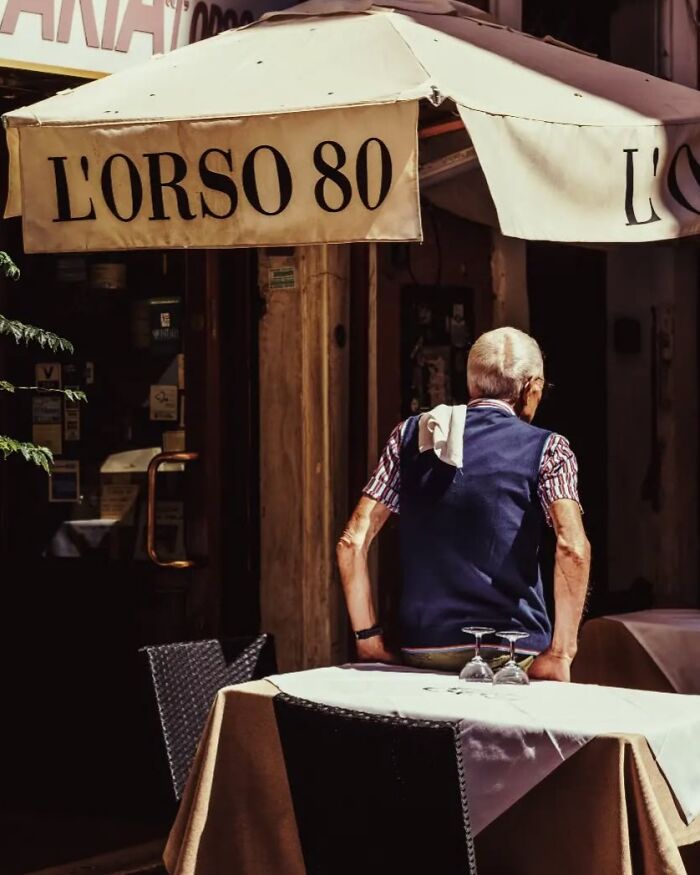 Elderly man sitting at a table under an awning in a street photo by Greek photographer Marina Nota.