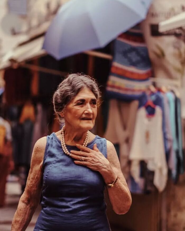 Elderly woman wearing pearls and a blue dress walking in a vibrant street scene captured in stunning street photos.