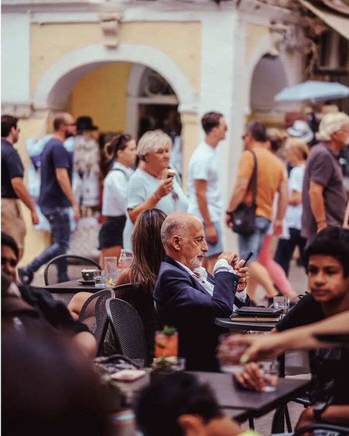 Older man using phone at outdoor café with blurred pedestrians walking by, a stunning street photo by Greek photographer Marina Nota