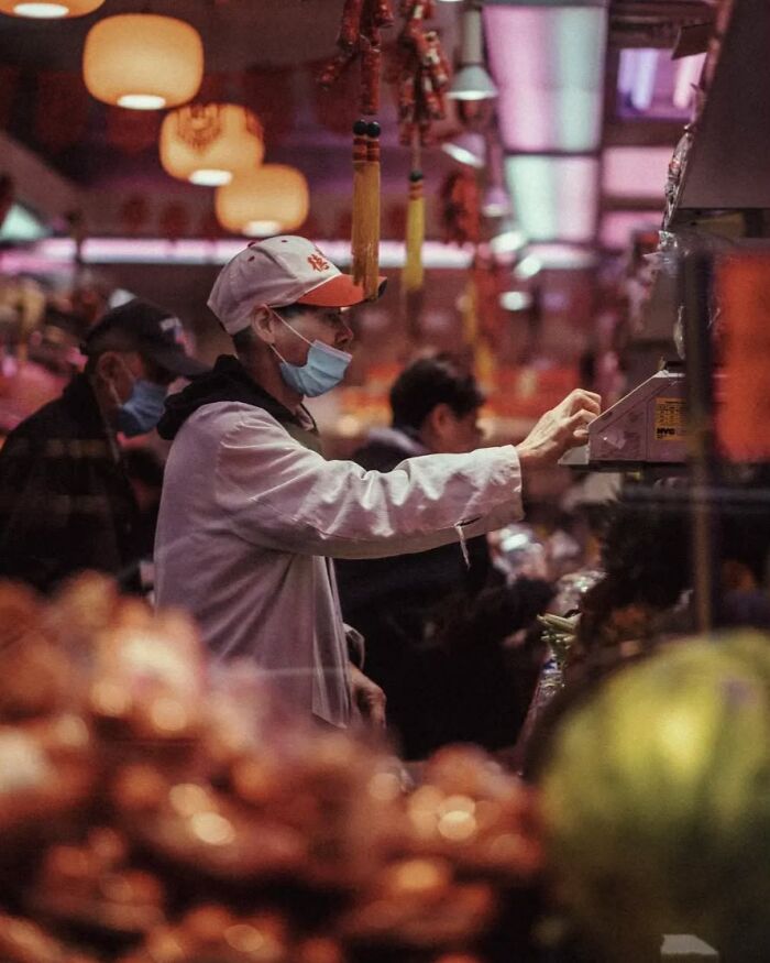 Person wearing a mask and cap interacting with a market scale, captured in stunning street photo by Greek photographer.