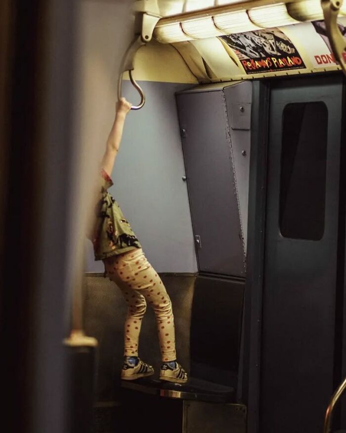 Child in polka dot pants hanging from a handrail inside a subway car, captured in a stunning street photo by Marina Nota.
