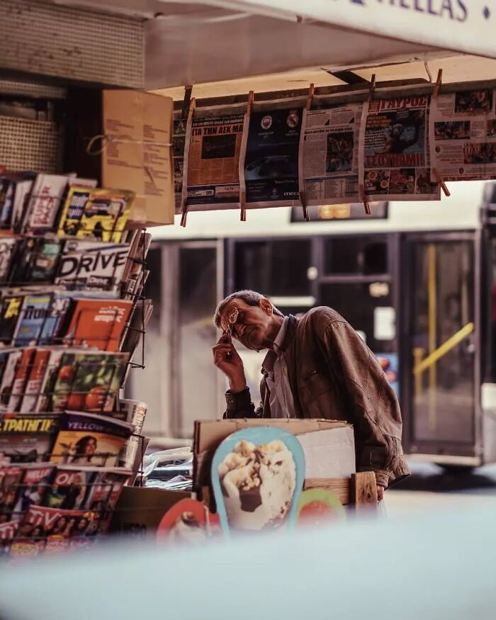 Man in a brown jacket reading newspapers at a busy urban street stand, captured in stunning street photos.