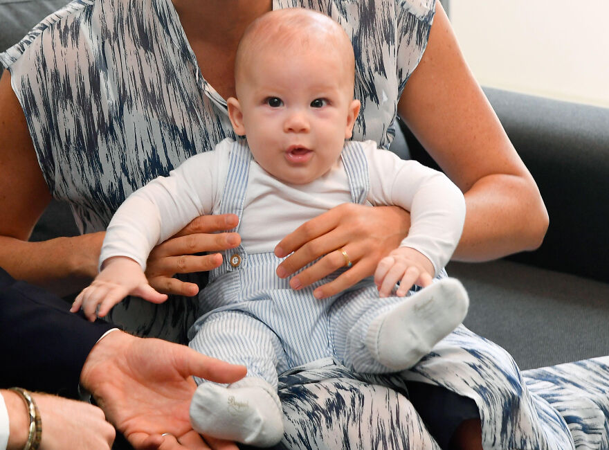 Baby held by a woman wearing patterned clothes, illustrating Meghan Markle and Prince Harry kids&rsquo; lives away from Buckingham Palace.