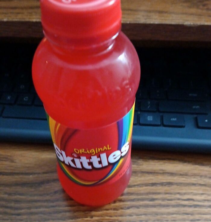 Red Skittles drink bottle on a wooden desk in front of a computer keyboard, showcasing a nonsensical product.