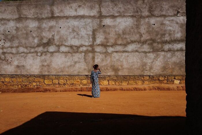 Child of the world standing alone on a sunlit dirt ground against a textured stone and plaster wall in powerful photograph