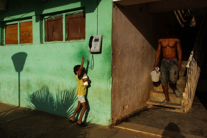 Child playing near a green wall holding a doll while a shirtless man descends stairs in warm sunlight in a powerful photograph