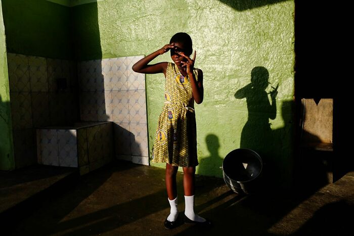 Child from powerful photographs by Andrea Torrei, posing in patterned dress with dramatic shadow on green wall background.