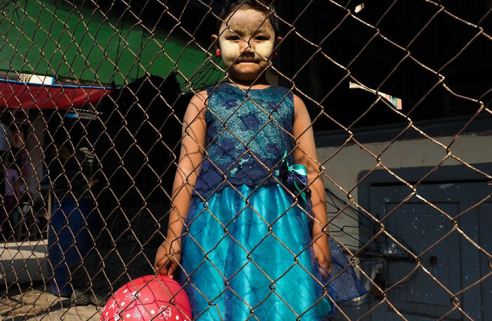 Young girl in a blue dress holding a pink ball behind a chain-link fence in powerful children of the world photograph