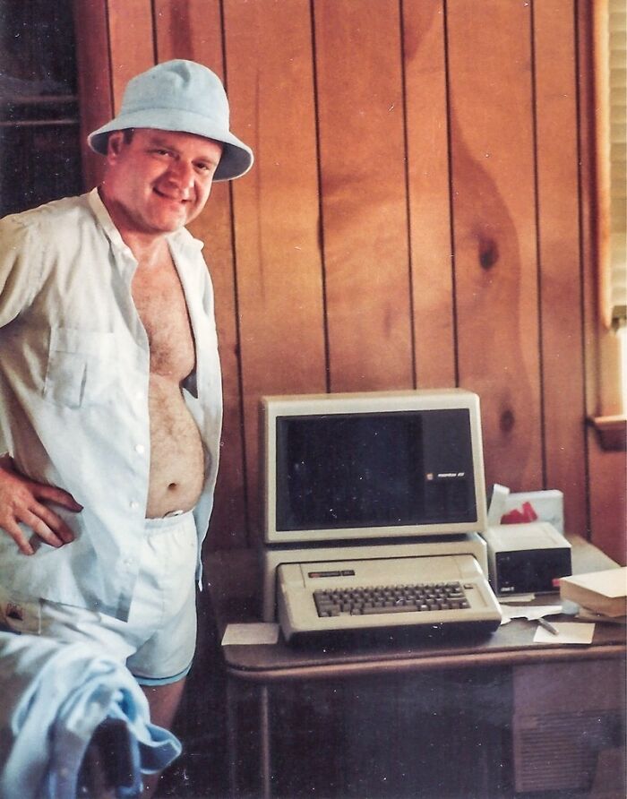Man in casual 80s vacation outfit standing next to a vintage computer inside a wood-paneled room, vintage vacation pics.