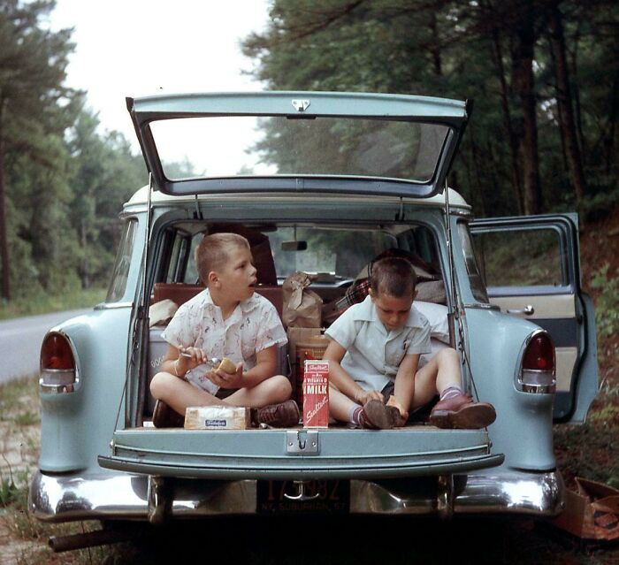 Two boys eating snacks from the trunk of an old car during a vintage vacation before cell phones had cameras