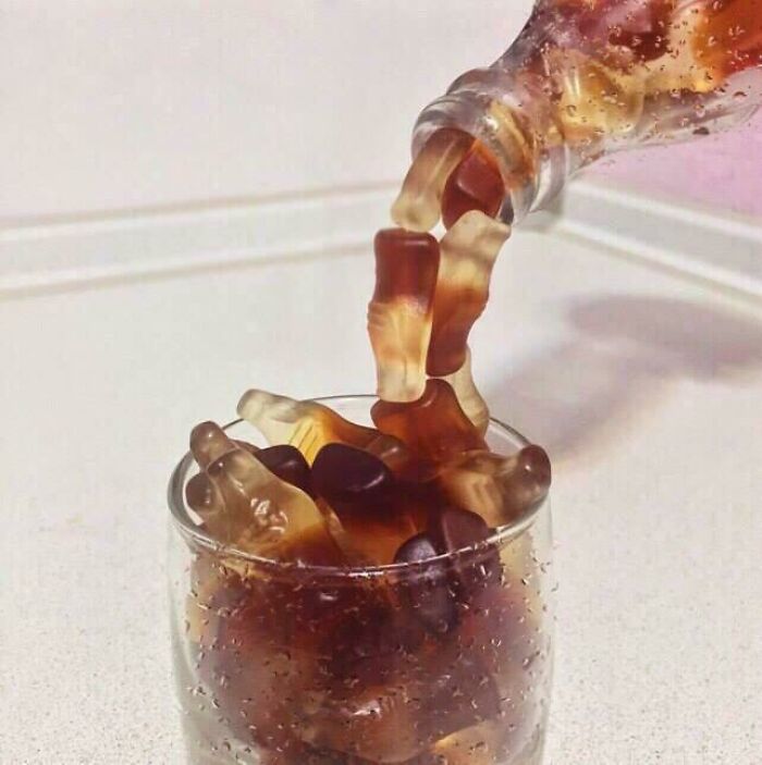 Glass filled with gummy cola bottles pouring from a soda bottle in a weird and blessed cursed unusual photo.