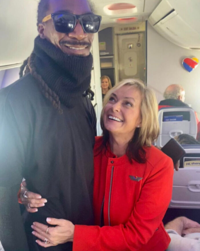 Man wearing sunglasses and turtleneck posing with smiling woman in red jacket on airplane, celebrity lookalike moment.