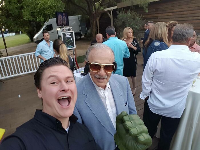 Man taking a selfie with an older man wearing sunglasses and a Hulk fist at a casual outdoor gathering with others nearby.