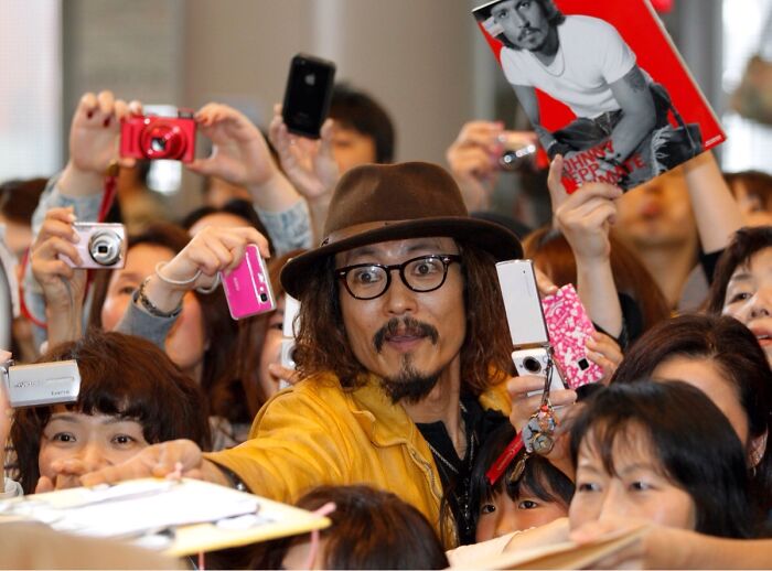 Man in a hat and glasses surrounded by fans taking photos, capturing a moment with a celebrity look-alike.