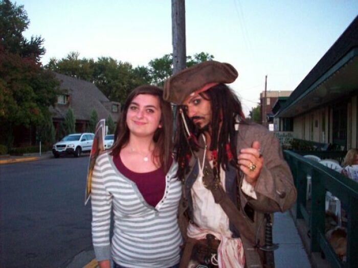 A woman posing with a Johnny Derp celebrity lookalike dressed as a pirate on a suburban street at dusk.