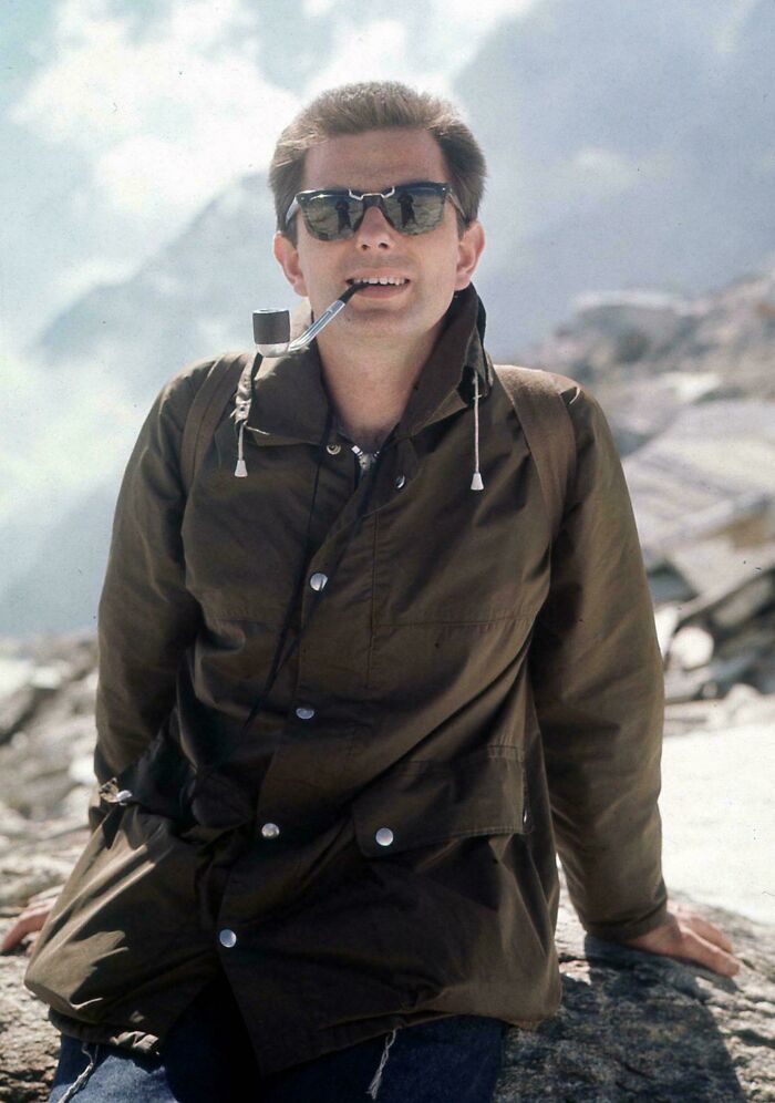 Man with pipe and sunglasses sitting on a rock during vacation in a scenic mountain setting before cell phone cameras.