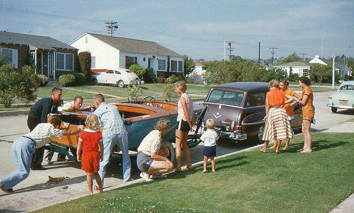 Family loading wooden boat in driveway during vintage vacation before cell phones had cameras in suburban neighborhood.