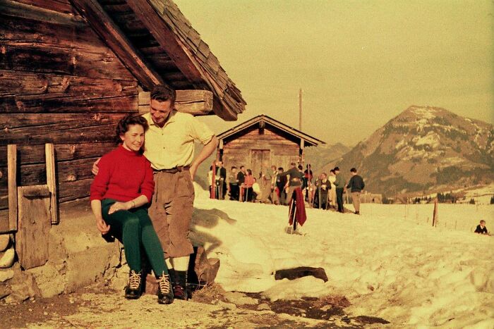 Vintage vacation pic of a couple outside a wooden cabin with snow and mountains in the background from before cell phones had cameras