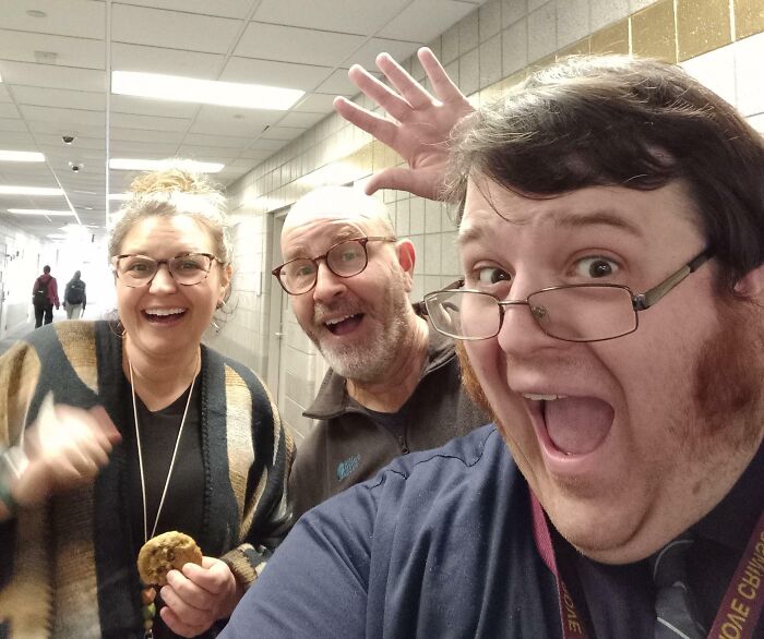 Three funny teachers smiling and posing for a playful selfie in a school hallway showcasing their sense of humor.
