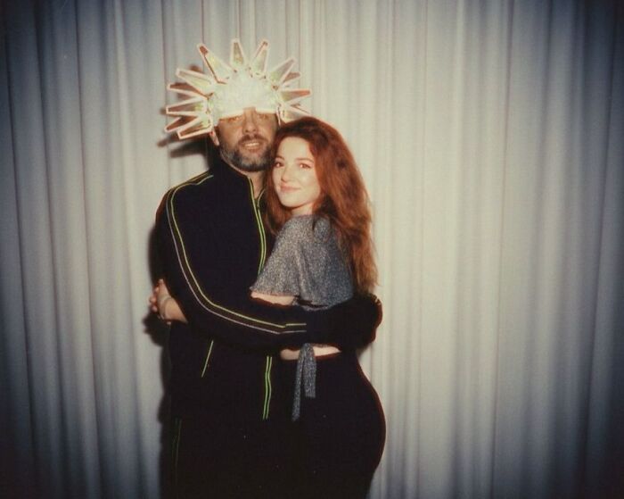 A man wearing a unique hat hugs a smiling woman, a funny celebrity moment shared and laughed at online.