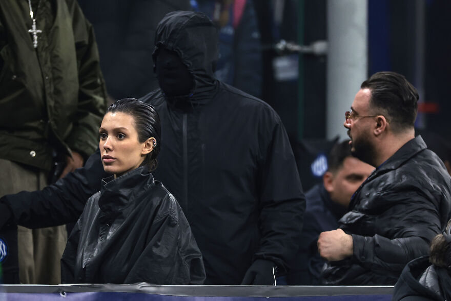 Bianca Censori wearing a dark jacket sitting among people, highlighting her role in Kanye West&rsquo;s growing empire and net worth.