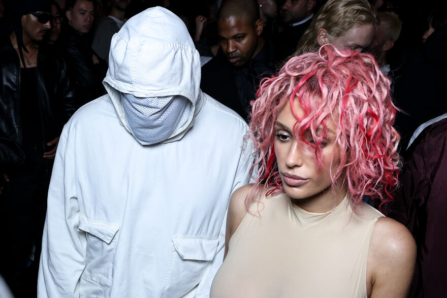 Bianca Censori with pink hair and West wearing a white hooded jacket at an event in a dark crowded setting.
