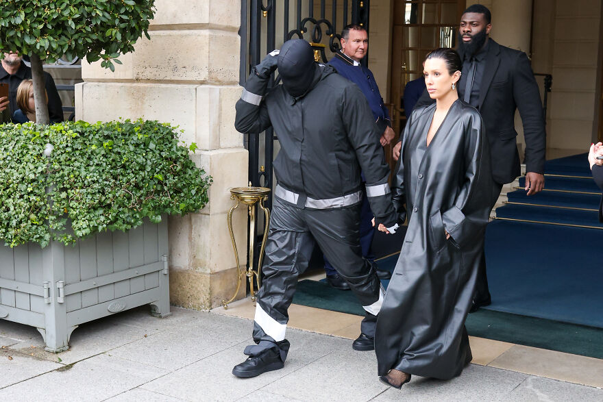 Bianca Censori wearing a long black coat walking with Kanye West and security outside a building.