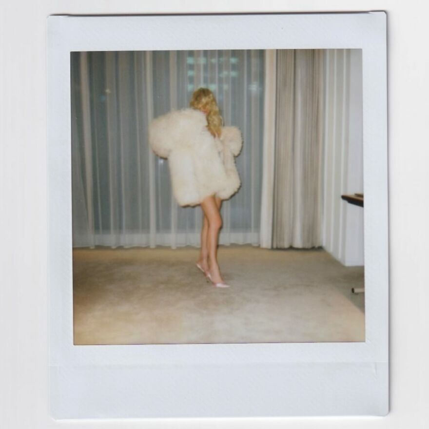 Bianca Censori wearing a white fur coat and heels standing in a room with curtains, related to Bianca Censori net worth and West empire.