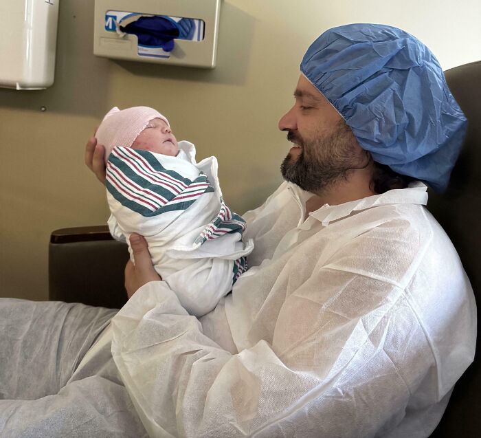 Father in hospital attire smiling while holding his newborn baby wrapped in a blanket, showcasing wholesome parent moments.