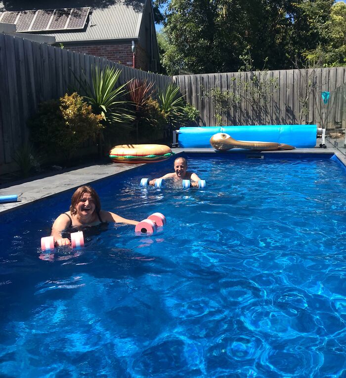 Parents having fun in a backyard pool with kids, using colorful foam dumbbells on a sunny day outdoors.