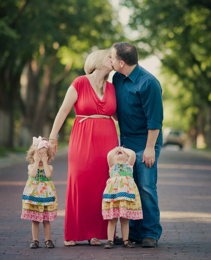 Parents sharing a funny and wholesome moment kissing while their kids playfully cover their eyes outdoors.