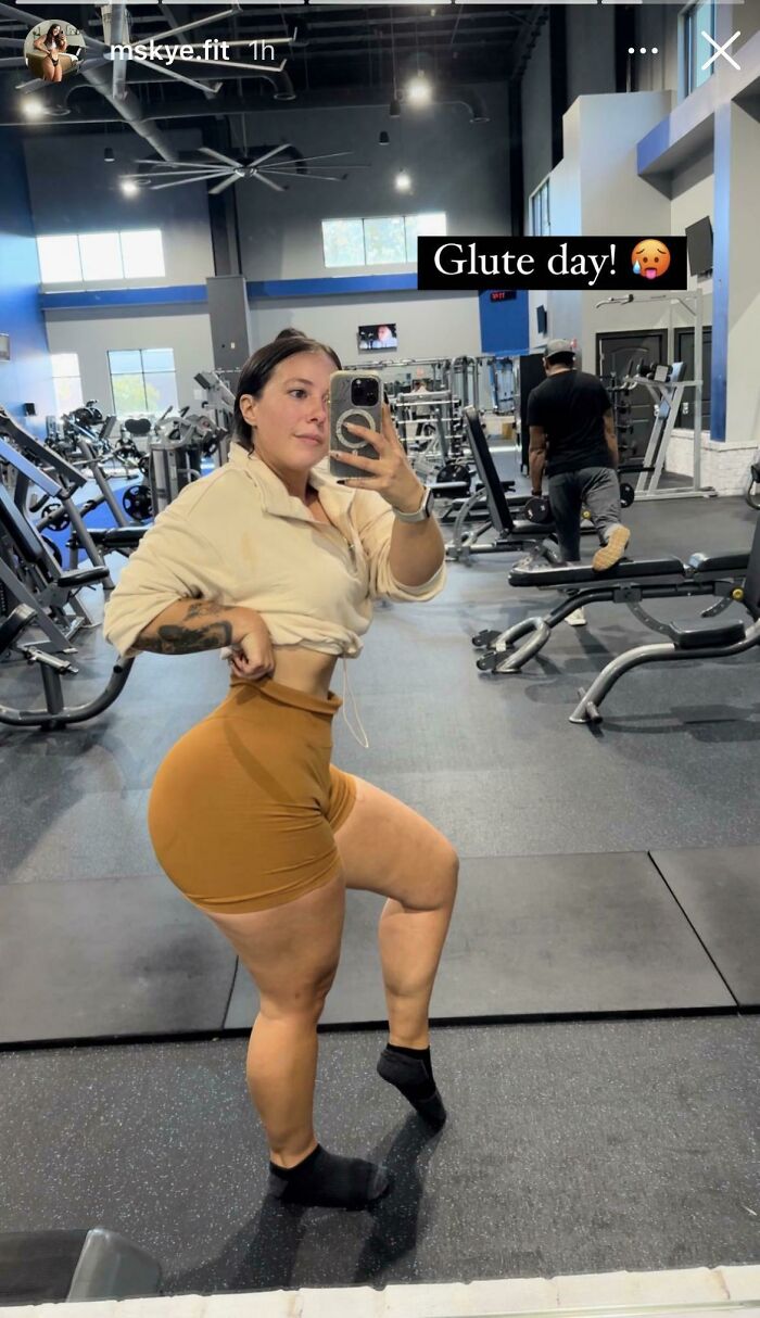 Woman taking a gym selfie in shorts with a focus on glute workout, surrounded by fitness equipment in the background