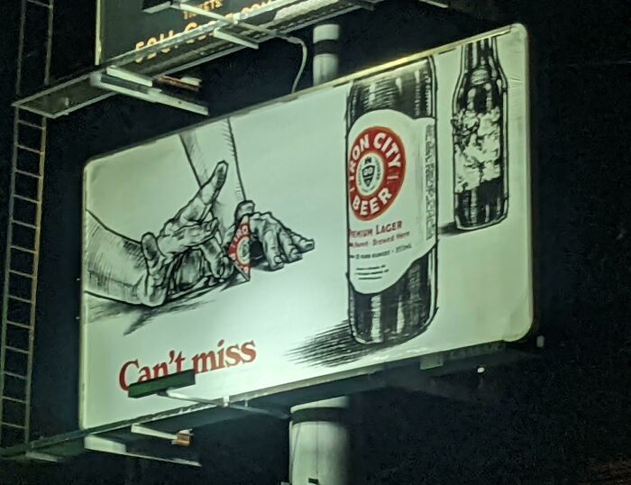 Funny and weird billboard at night showing hands flipping a small beer bottle with large beer bottles in background