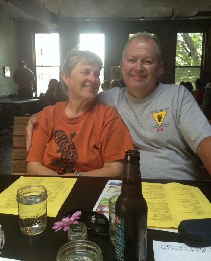 Smiling parents sitting close together at a restaurant table with drinks and menus, capturing a funny and wholesome moment.