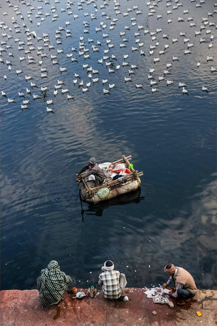Man on a small boat and three people sitting by the water in India, surrounded by numerous floating birds.
