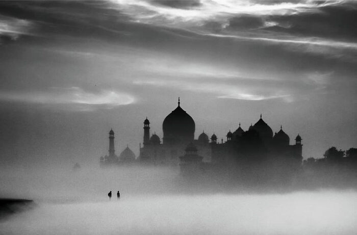 Black and white photo capturing everyday life in India with two people walking near the iconic Taj Mahal in misty conditions.